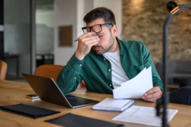 Stressed man sitting at a desk, holding papers in one hand while working on a laptop, with a worried expression on his face.